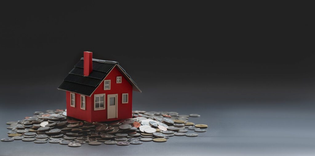Malý červený model domu stojí na rozsypanej hromade mincí na tmavom, rozmazanom pozadí a symbolizuje investíciu do domu, financie na nehnuteľnosti alebo význam úveru na bývanie.