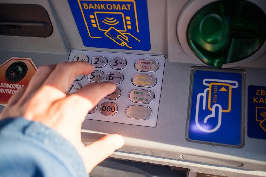 Ruka stláčajúca tlačidlá na klávesnici bankomatu s nápismi a symbolmi pre platbu kartou a bezkontaktnú platbu a textové nápisy v poľštine viditeľné v okolí bankomatu.