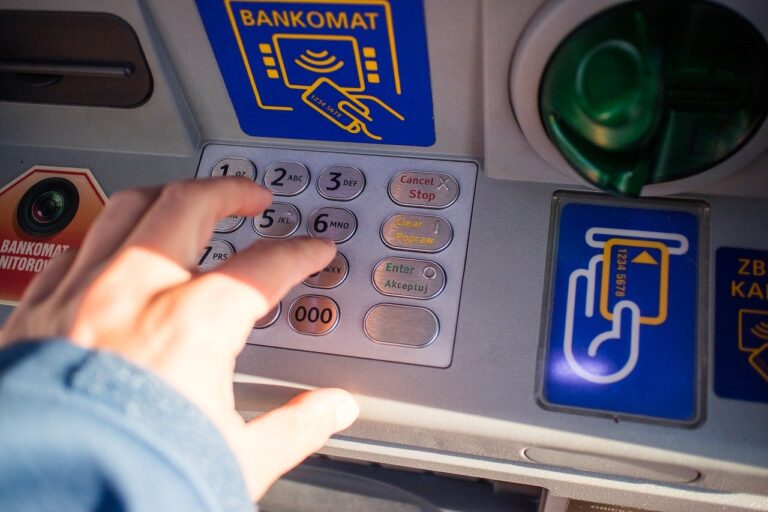 Ruka stláčajúca tlačidlá na klávesnici bankomatu s nápismi a symbolmi pre platbu kartou a bezkontaktnú platbu a textové nápisy v poľštine viditeľné v okolí bankomatu.