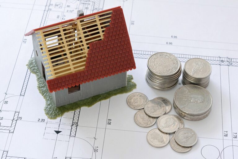 Model domu vo výstavbe stojí vedľa niekoľkých stohov a hromádok mincí, ktoré sú umiestnené na architektonických plánoch a symbolizujú splácanie úveru počas celého procesu výstavby.