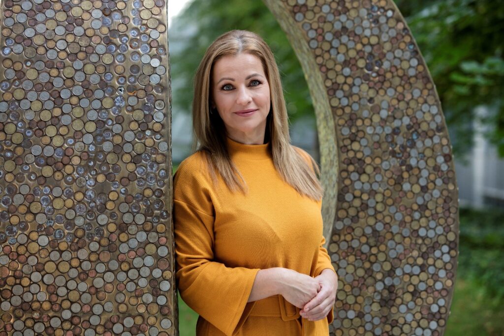 Žena v horčicovožltých šatách stojí vonku, usmieva sa a pózuje so zopnutými rukami pred veľkou kruhovou kovovou plastikou pokrytou vzormi mincí, ktoré odrážajú História kremnickej mincovne. V pozadí je vidieť zelené lístie.
