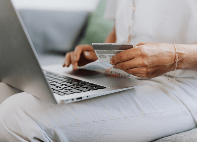 Osoba sedí s notebookom na kolenách, v jednej ruke drží kreditnú kartu a druhou píše na klávesnici, čo naznačuje online nakupovanie alebo platba kartou.