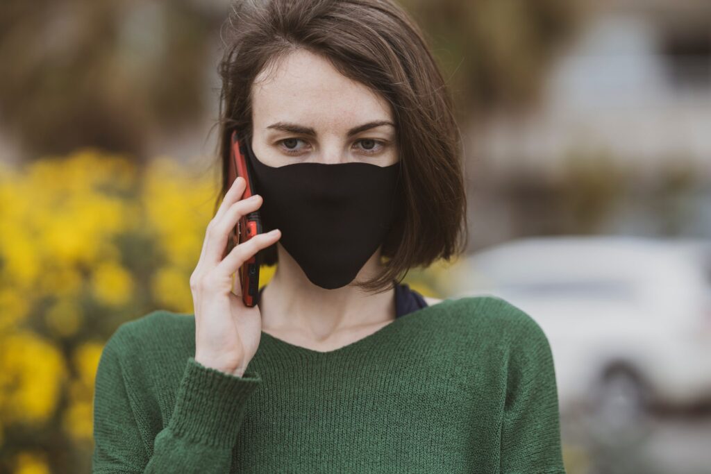 Žena s hnedými vlasmi v čiernej maske na tvári a zelenom svetri hovorí na svojom smartfóne vonku, v pozadí sú žlté kvety a rozmazané biele auto.