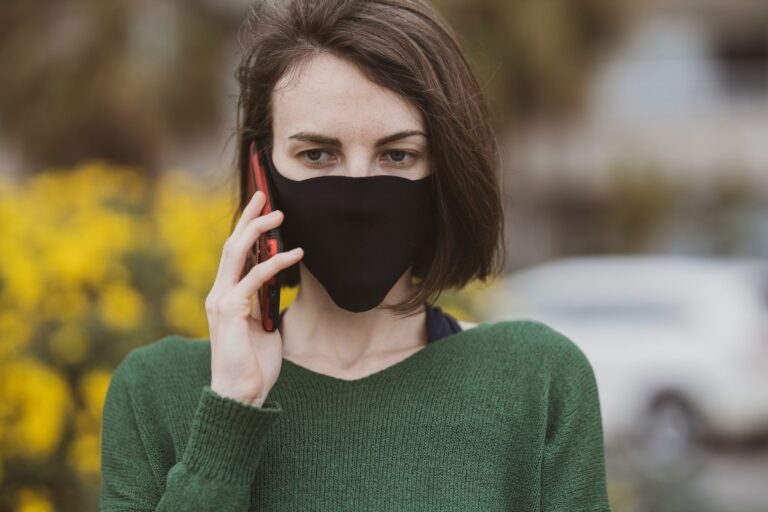 Žena s hnedými vlasmi v čiernej maske na tvári a zelenom svetri hovorí na svojom smartfóne vonku, v pozadí sú žlté kvety a rozmazané biele auto.