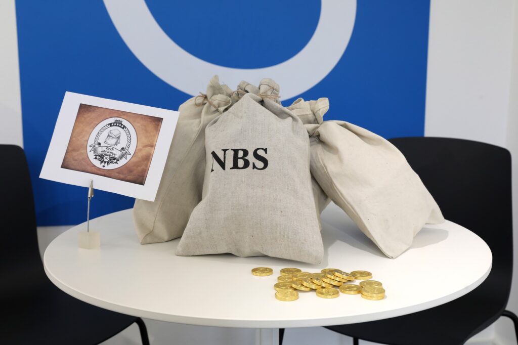 Tri plátenné tašky s nápisom NBS ležia na bielom stole s rozsypanými zlatými mincami a malou tabuľkou s ilustrovanou pečaťou; vidno dve čierne stoličky a modré a biele pozadie.