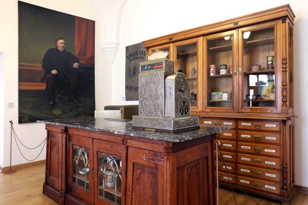 Interiér historickej lekárničky s dreveným pultom so zdobenou pokladňou, vitrínou plnou pohárov a dóz a veľkým portrétom muža na stene.