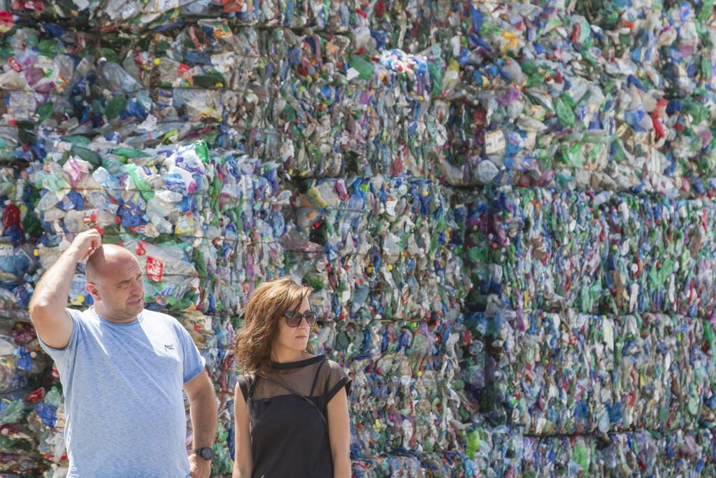 Dvaja ľudia stoja pred veľkými kopami stlačených plastových fliaš a iných recyklovateľných materiálov, muž sa škrabe na hlave a žena má slnečné okuliare. Scéna naznačuje, že ide o zariadenie na recykláciu alebo nakladanie s odpadom.