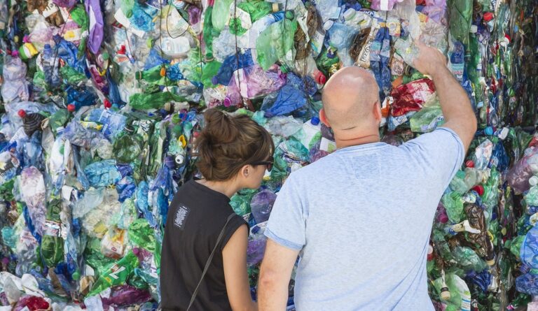 Dvaja ľudia, žena a holohlavý muž, stoja pred veľkou stenou zo stlačených plastových fliaš a nádob a skúmajú a diskutujú o pestrej hromade recyklovateľného odpadu.