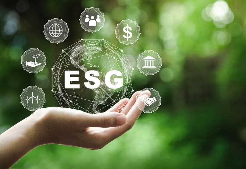 Ruka drží digitálny glóbus s nápisom ESG v strede, obklopený ikonami predstavujúcimi udržateľnosť, financie, komunitu, správu vecí verejných a obnoviteľnú energiu na rozmazanom zelenom pozadí.