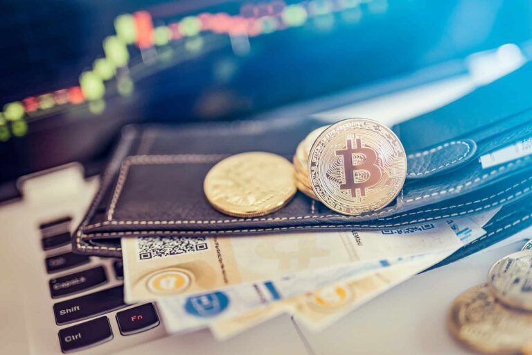 Detailný záber na klávesnicu notebooku s mincami Bitcoin, papierovou menou a peňaženkou. V pozadí je viditeľný digitálny akciový graf, ktorý naznačuje obchodovanie s kryptomenami alebo investovanie.