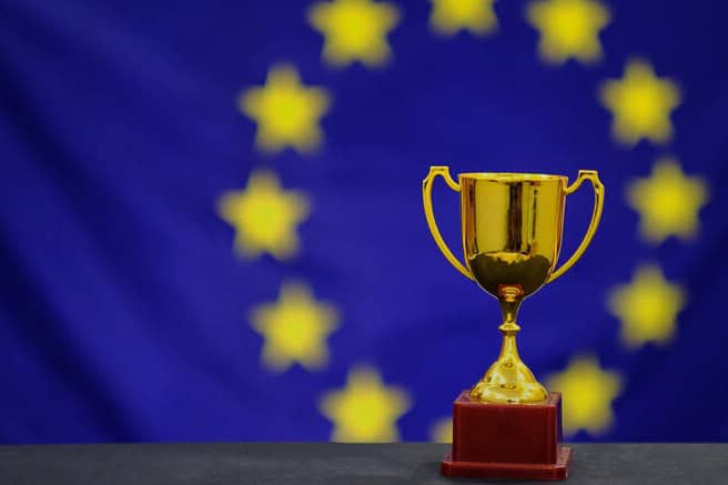 Zlatá trofej na červenom podklade stojí pred rozmazanou vlajkou Európskej únie so žltými hviezdami na modrom pozadí.