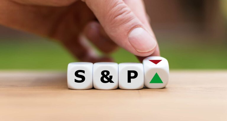 Ruka otáča kockami na drevenom povrchu a ukazuje písmená "S & P" a zelený trojuholník smerujúci nahor vedľa červeného trojuholníka smerujúceho nadol, čo symbolizuje zmeny v indexe S&P.