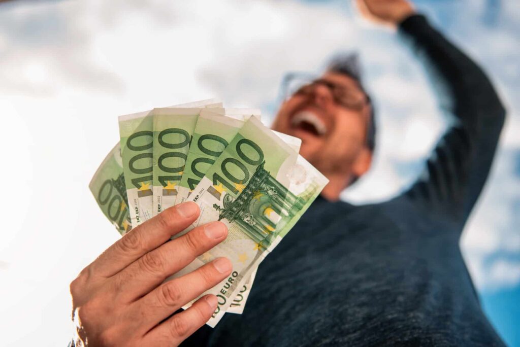 Osoba, ktorá drží v jednej ruke niekoľko 100-eurových bankoviek, usmieva sa a druhú ruku dvíha na oslavu na pozadí modrej oblohy.