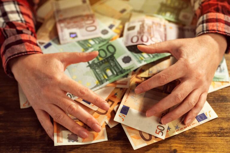Osoba v červenej kockovanej košeli rozprestiera ruky nad kopou rôznych eurobankoviek na drevenom povrchu.