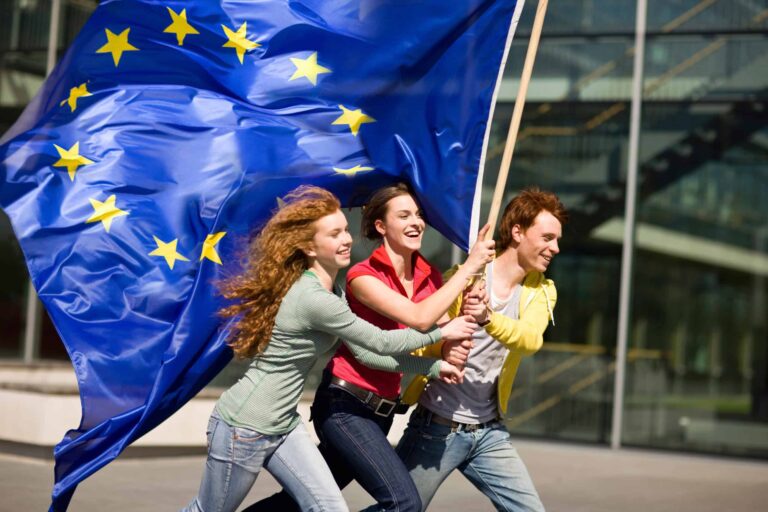 Traja usmievaví mladí ľudia bežia vonku a držia veľkú vlajku Európskej únie, v pozadí sú moderné sklenené budovy. Vlajka sa za nimi vlní a zobrazuje žlté hviezdy na modrom poli.