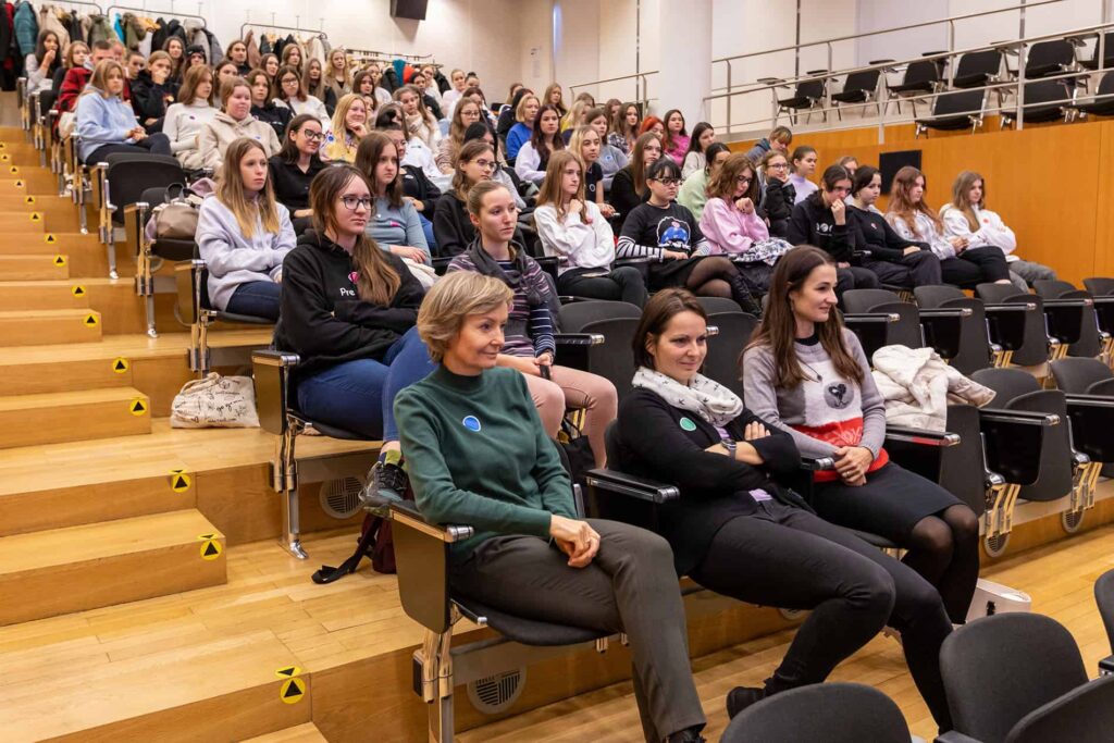 Veľká skupina ľudí, väčšinou mladých žien, sedí v prednáškovej sále so sedadlami ako na štadióne a pozorne počúva prezentáciu alebo prednášku. Miestnosť je svetlá, s drevenou podlahou a modernými čiernymi stoličkami.