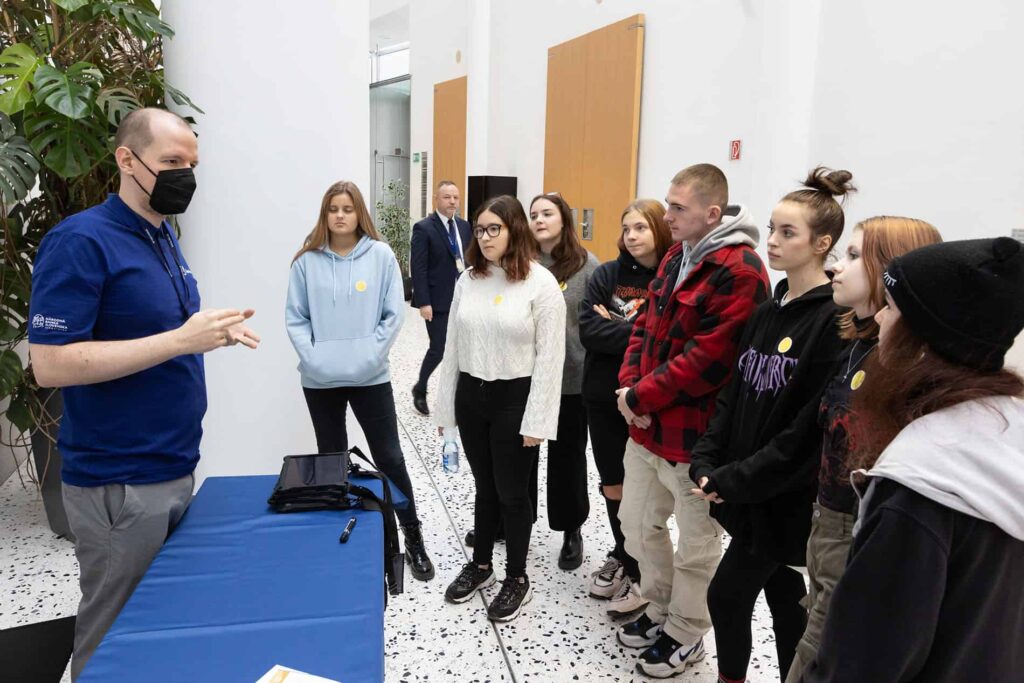 Muž v modrej košeli a čiernej maske sa prihovára skupine mladých dospelých, ktorí stoja pred ním v interiéri; skupina pozorne počúva pri stole s písacím stolíkom a perom.