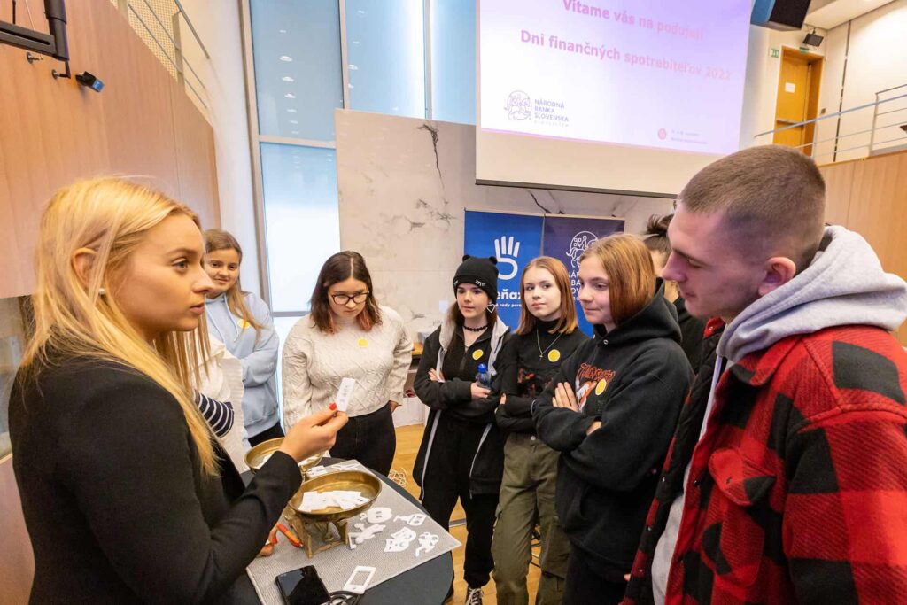 Žena niečo vysvetľuje, pričom drží karty skupine mladých ľudí zhromaždených okolo stola v učebni alebo seminárnej miestnosti, pričom v pozadí sa premieta prezentácia.