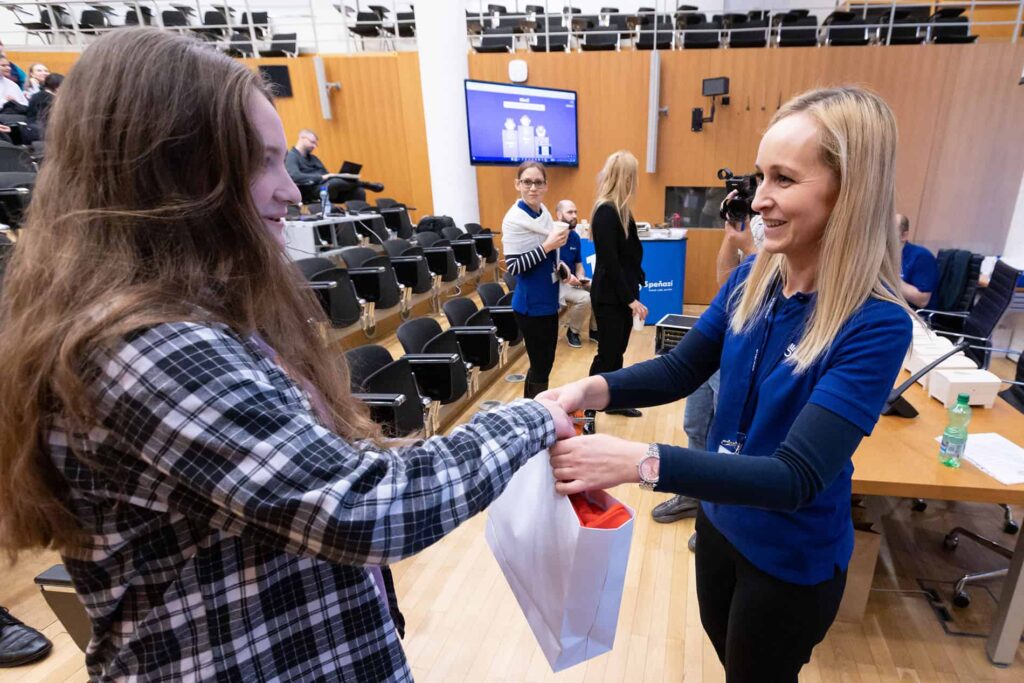 Žena v modrej košeli podáva darčekovú tašku mladému človeku v kockovanej košeli v prednáškovej sále, kde sedia ďalší ľudia a v pozadí je vidieť veľkú obrazovku.