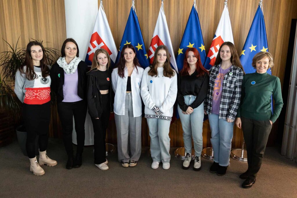 Sedem mladých žien a jedna staršia žena stoja v rade v interiéri a usmievajú sa do kamery. Za nimi sú slovenské vlajky a vlajky Európskej únie. Prostredie pôsobí formálne, v pozadí je drevom obložená stena.