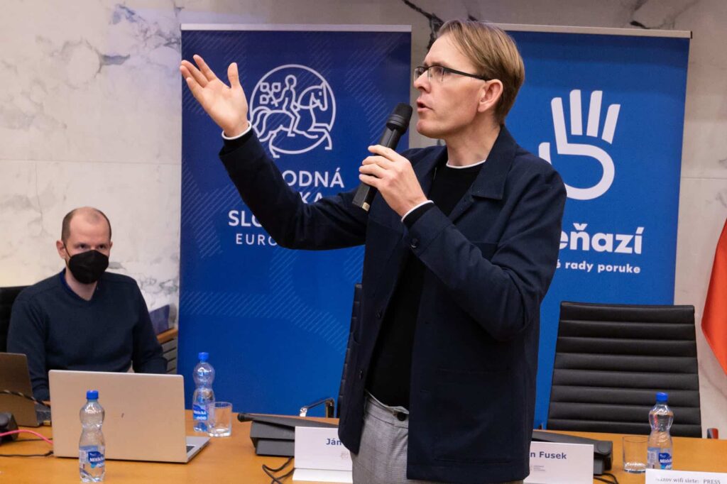 Muž stojí a hovorí do mikrofónu so zdvihnutou rukou, zatiaľ čo iný muž sedí za stolom s notebookmi a maskou na tvári. V pozadí sú modré transparenty so slovenským textom.