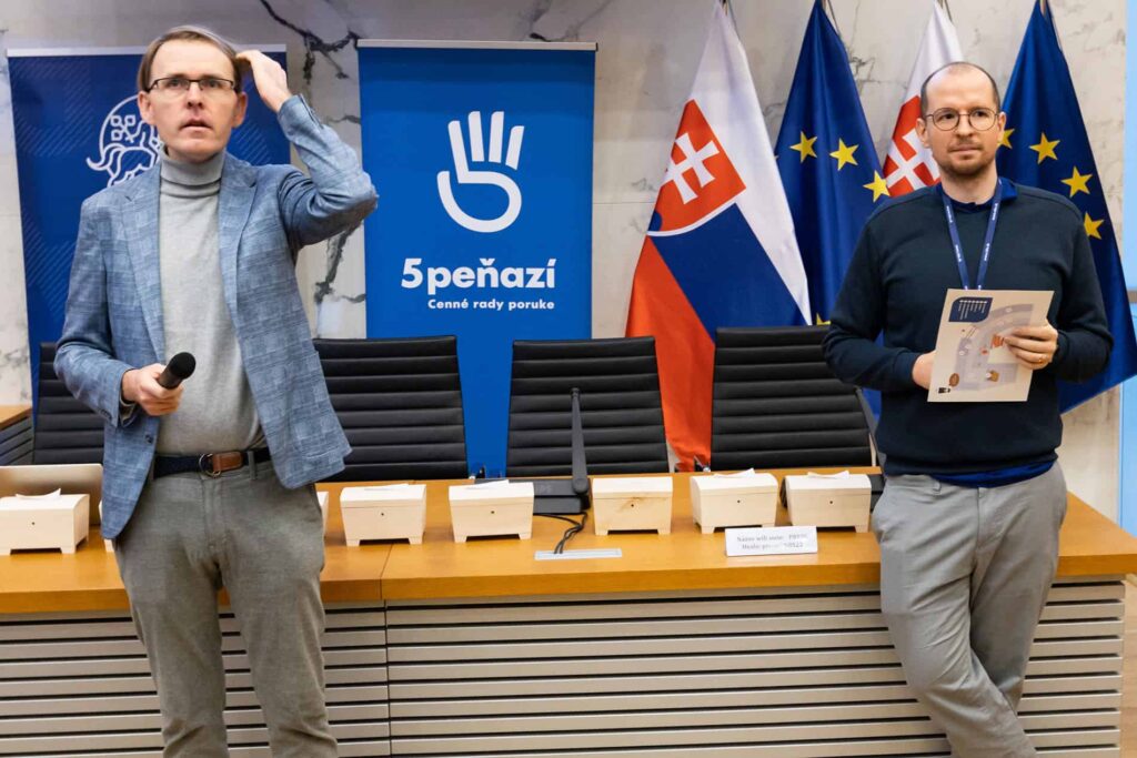 Dvaja muži stoja pred konferenčným stolom s mikrofónmi a malými škatuľkami. Za nimi sú vlajky Slovenska a EÚ a modrý transparent s textom "5PEŇAZÍ". Jeden muž drží mikrofón, druhý drží papiere.