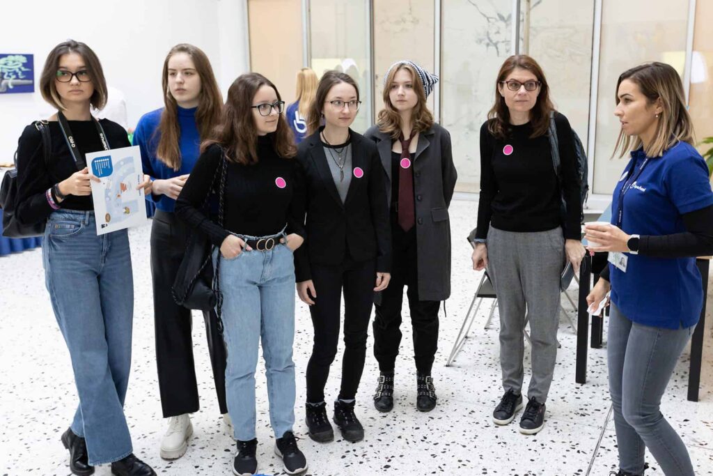 Sedem žien stojí vo vnútri v skupine, šesť v čiernom a jedna v modrom s visačkou s menom. Väčšina z nich má na tričkách ružové nálepky. Jedna z nich drží papier a zdá sa, že počúvajú alebo sa pripravujú na nejakú udalosť.
