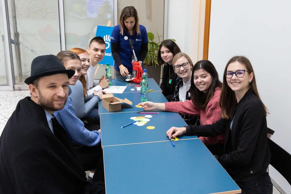 Skupina mladých dospelých sedí okolo modrého stola, usmieva sa do kamery a pred sebou má papiere, farebné kruhy a písacie potreby. Za stolom stojí žena a pomáha pri činnosti.