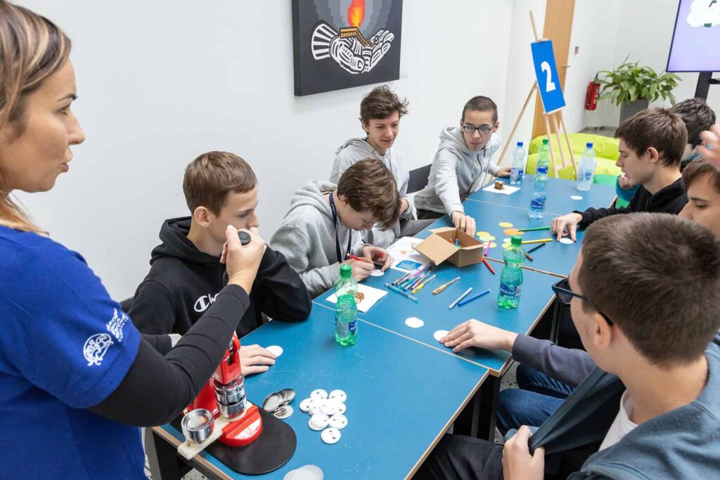 Skupina chlapcov sedí pri modrých stoloch a pracuje na remeselnom alebo vedeckom projekte s gombíkmi, fixkami a farebnými ceruzkami, zatiaľ čo žena stojí neďaleko a dohliada na stroj na výrobu gombíkov.