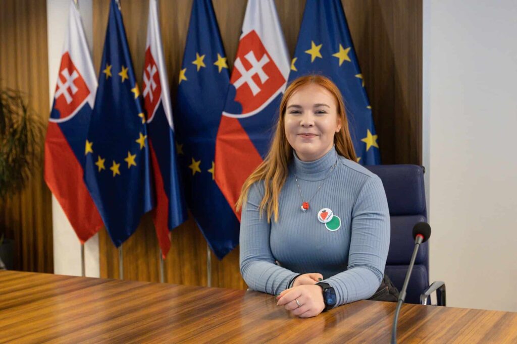 Žena s dlhými ryšavými vlasmi sedí za konferenčným stolom a usmieva sa pred vlajkami Európskej únie a Slovenska. Na sebe má svetlomodrý rolák a na košeli pripnuté odznaky.