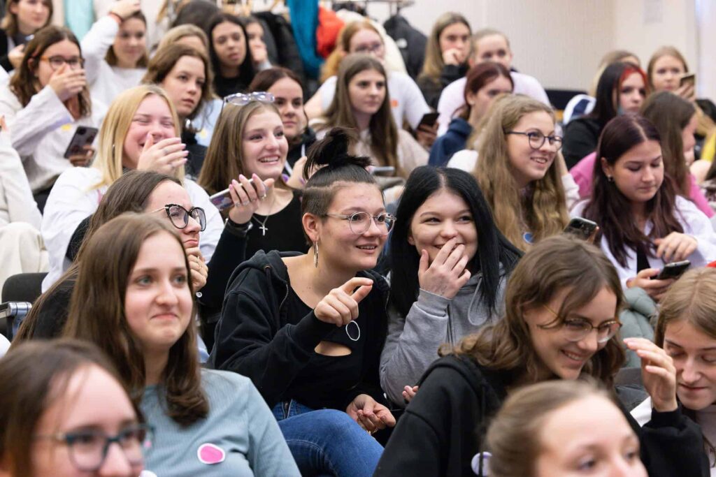 Skupina mladých žien sedí tesne vedľa seba v interiéri, mnohé sa usmievajú a smejú, niektoré držia smartfóny a majú okuliare, zdá sa, že sa tešia zo spoločnej udalosti alebo prezentácie.