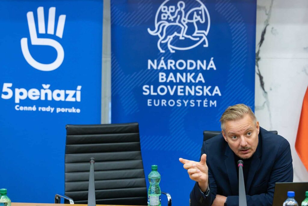 Muž v tmavom obleku hovorí a gestikuluje pri stole s fľašami s vodou a mikrofónmi, sedí medzi dvoma transparentmi, medzi ktorými je aj transparent Národnej banky Slovenska.