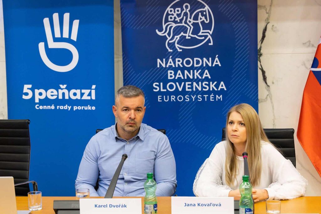 Dvaja ľudia, muž a žena, sedia za stolom s mikrofónmi a tabuľkami s menami pred kulisou Národnej banky Slovenska. Na stole sú fľaše s vodou. Žena vyzerá sústredene, muž sa tvári vážne.
