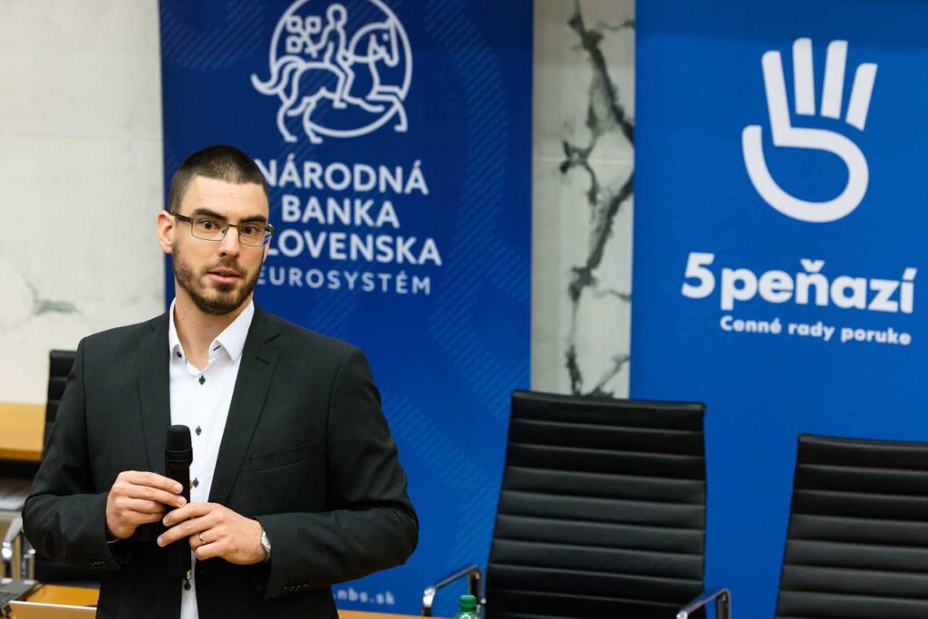 Muž v čiernom obleku a okuliaroch drží mikrofón a stojí pred transparentmi so slovenskými textami a logami, medzi ktorými sú Národná banka Slovenska a 5PEŇAZÍ. Za ním je vidieť prázdne stoličky.