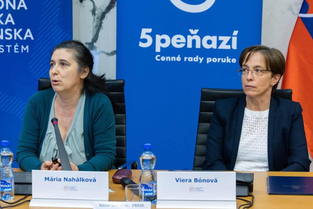 Dve ženy sedia pri stole s mikrofónmi a fľašami s vodou. Žena naľavo hovorí, zatiaľ čo žena napravo počúva. Na menovkách je napísané Mária Nahálková a Viera Bónová. V pozadí sú modré a oranžové transparenty.