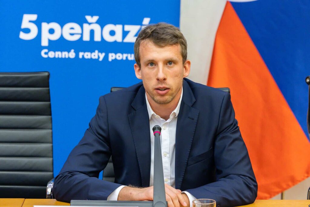 Muž v námorníckom obleku hovorí do mikrofónu pri konferenčnom stole. Za ním je modrá tabuľa so slovenským textom a čiastočná slovenská vlajka.