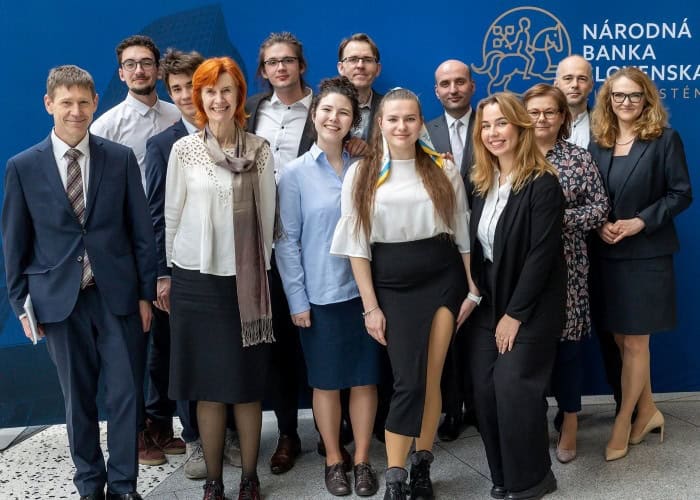 Skupina trinástich profesionálne oblečených ľudí stojí a usmieva sa pred modrou stenou, v pozadí vidno logo Národnej banky Slovenska.