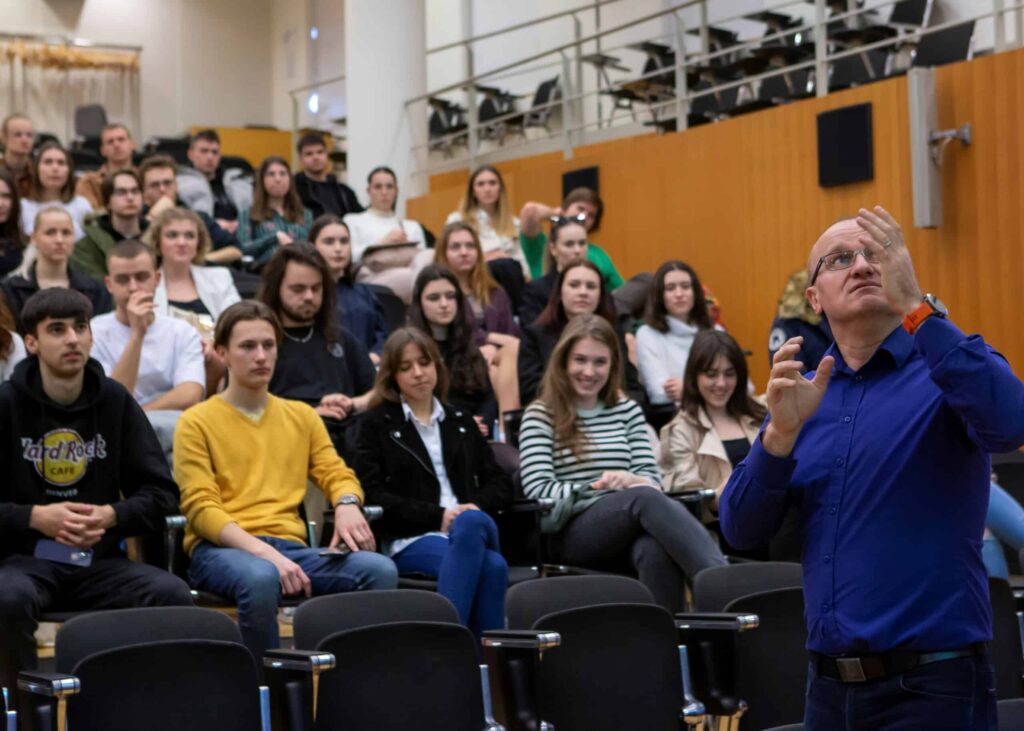 Muž gestikuluje, kým hovorí k skupine študentov sediacich v prednáškovej sále s drevenými stenami a radmi stoličiek. Študenti pozorne sledujú a počúvajú.