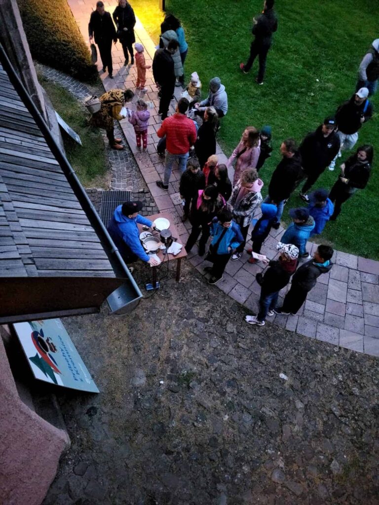 Skupina ľudí vrátane dospelých a detí sa za súmraku zhromaždila vonku pri budove. Niektorí stoja v rade pri stole, pri ktorom dvaja ľudia zrejme podávajú jedlo alebo nápoje. Vidno trávu a chodníky.
