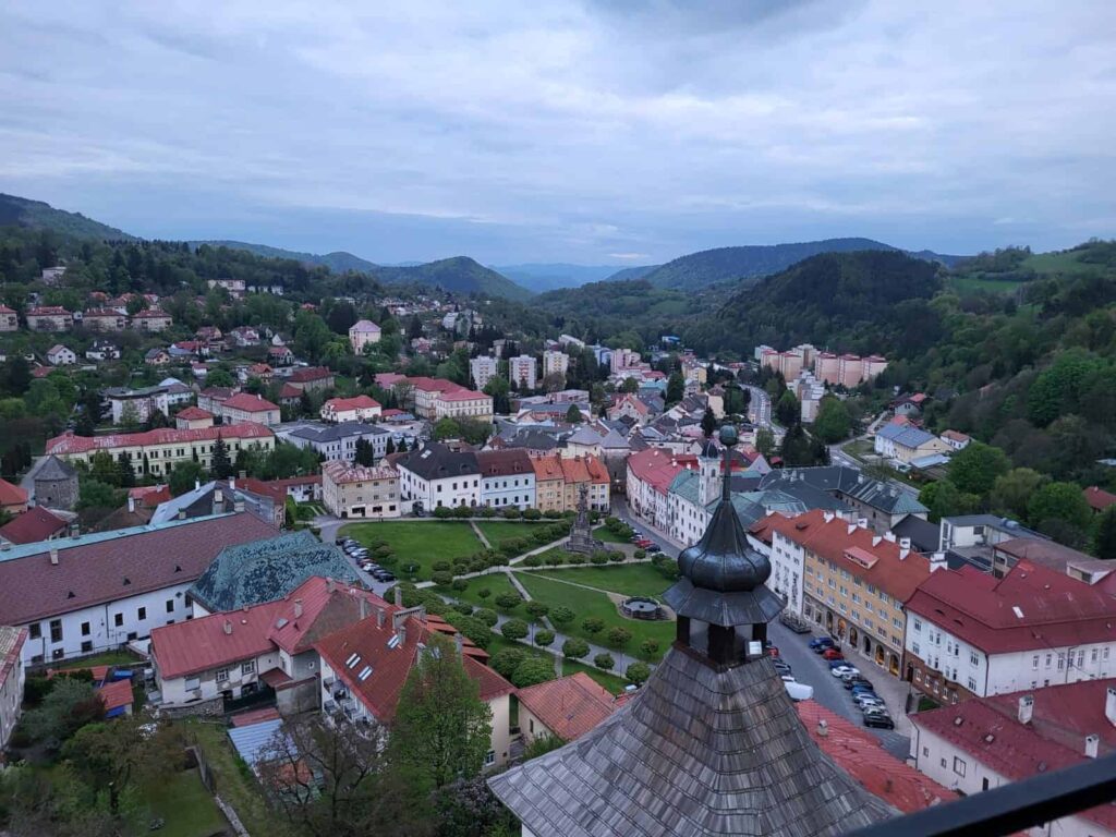 Malebný pohľad na mesto s červenými strechami, kľukatými uličkami, zelenými stromami a okolitými kopcami pod zamračenou oblohou. V popredí je viditeľná drevená kostolná veža.