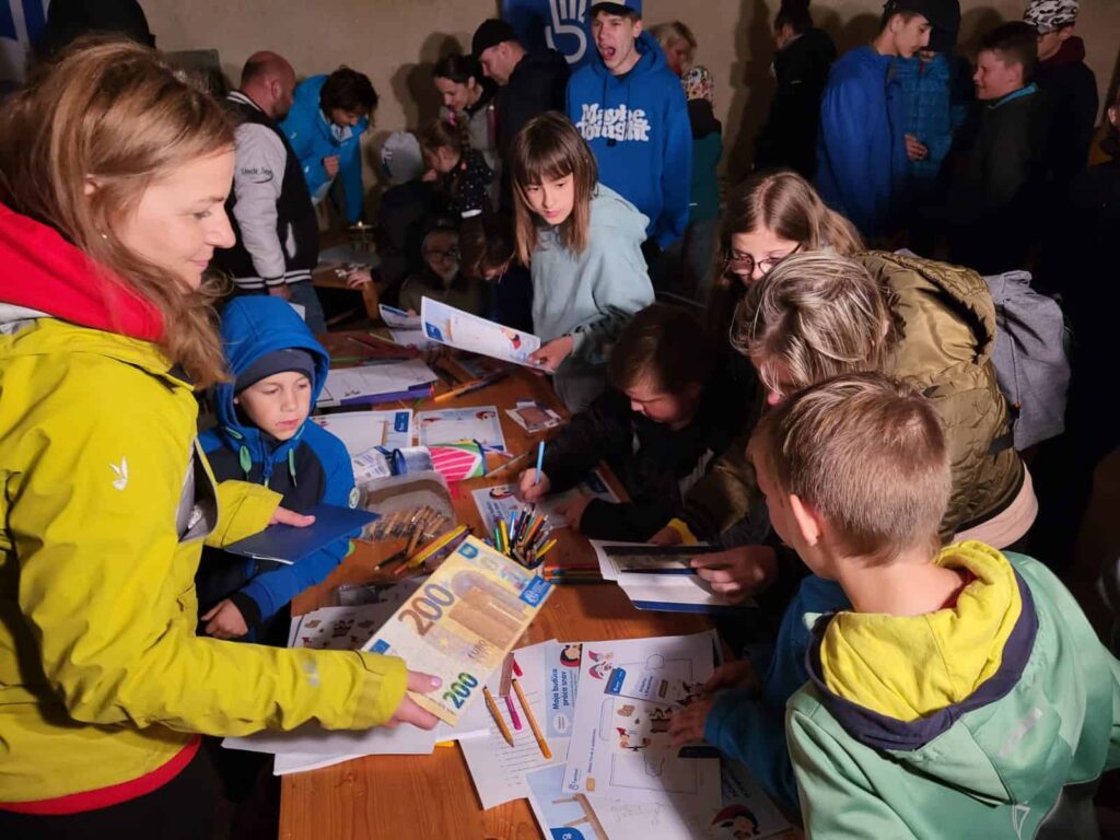 Skupina detí a dospelých sa na podujatí v interiéri zhromažďuje okolo stola s papiermi, ceruzkami, farbičkami a peniazmi na hranie a venuje sa tvorivým činnostiam.