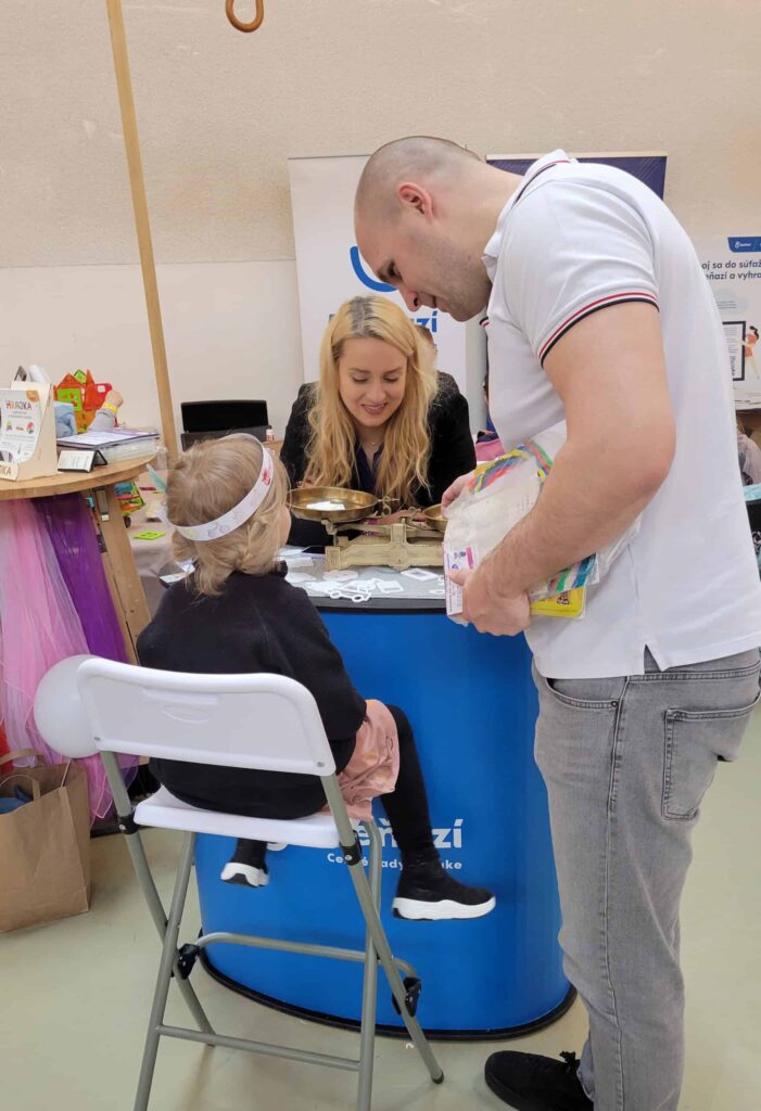Malé dieťa sedí na stoličke pri stánku, zatiaľ čo muž drží v ruke tovar a rozpráva sa so ženou za pultom. Žena sa usmieva a komunikuje s nimi. Stánok má modrý poťah na stole a informačné materiály.