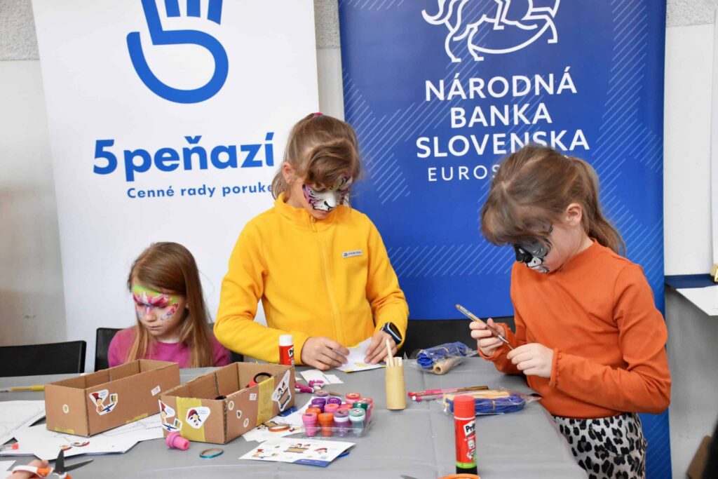Tri mladé dievčatá s farbami na tvár pracujú na výtvarných prácach pri stole so zásobami. Za nimi sú transparenty Národnej banky Slovenska a 5PEŇAZÍ. Na stole sú kartóny, lepiace tyčinky a fixky.