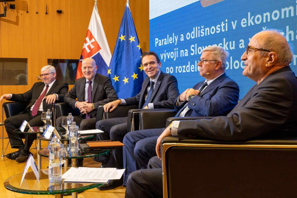 Päť mužov v oblekoch sedí na pódiu pred slovenskými vlajkami a vlajkami EÚ a zúčastňuje sa na panelovej diskusii. Za nimi je vidieť veľkú obrazovku so slovenským textom, na stole sú mikrofóny a fľaše s vodou.