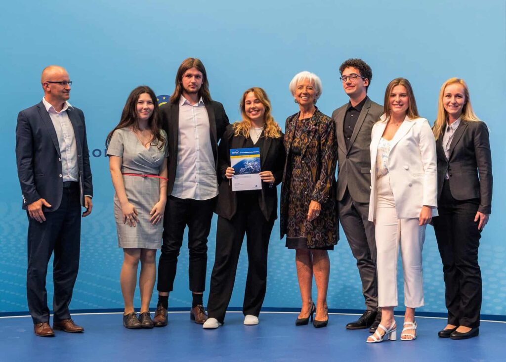 Na modrom pódiu stojí skupina siedmich ľudí, štyri ženy a traja muži. Jedna žena uprostred drží certifikát a usmieva sa. Všetci sú oblečení v obchodnom alebo formálnom oblečení a pózujú na skupinovú fotografiu.