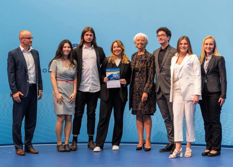 Na modrom pódiu stojí skupina siedmich ľudí, štyri ženy a traja muži. Jedna žena uprostred drží certifikát a usmieva sa. Všetci sú oblečení v obchodnom alebo formálnom oblečení a pózujú na skupinovú fotografiu.