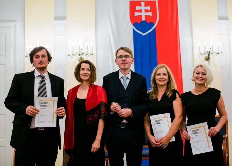 Päť ľudí v slávnostnom odeve stojí v interiéri, držia certifikáty a v pozadí je veľká slovenská vlajka a lustre. Na fotografii pózujú a usmievajú sa.