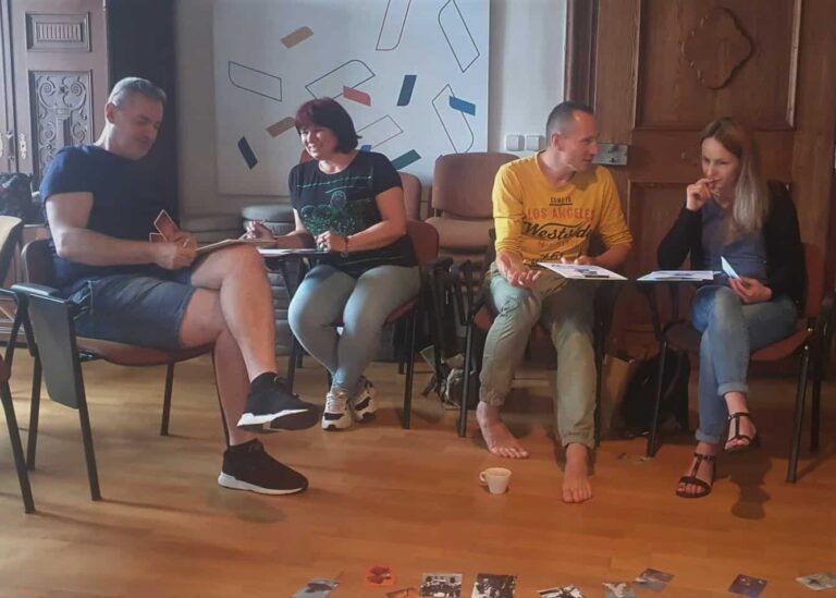 Štyria dospelí sedia vo dvojiciach na stoličkách v miestnosti a diskutujú a píšu. Na drevenej podlahe sú rozhádzané papiere a fotografie. Jedna osoba je bosá a atmosféra pôsobí neformálne a kolektívne.