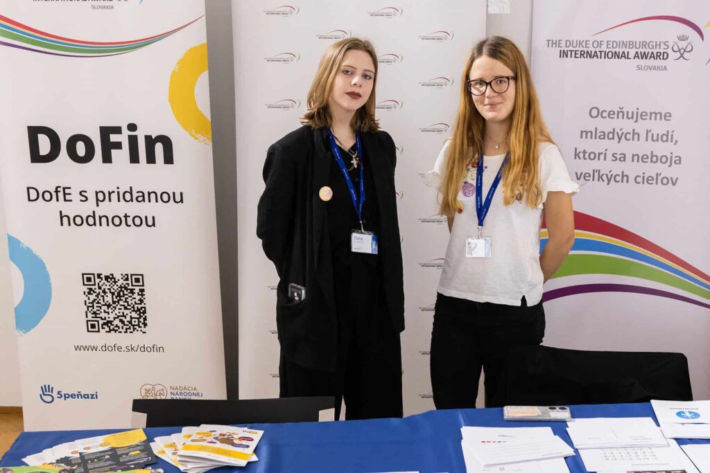 Dve mladé ženy stoja pri informačnom stánku pre Deň peňazí s transparentmi DoFin a Medzinárodnej ceny vojvodu z Edinburghu. Na stole sú vystavené brožúry, dokumenty a propagačné predmety. Obe ženy majú na sebe menovky.