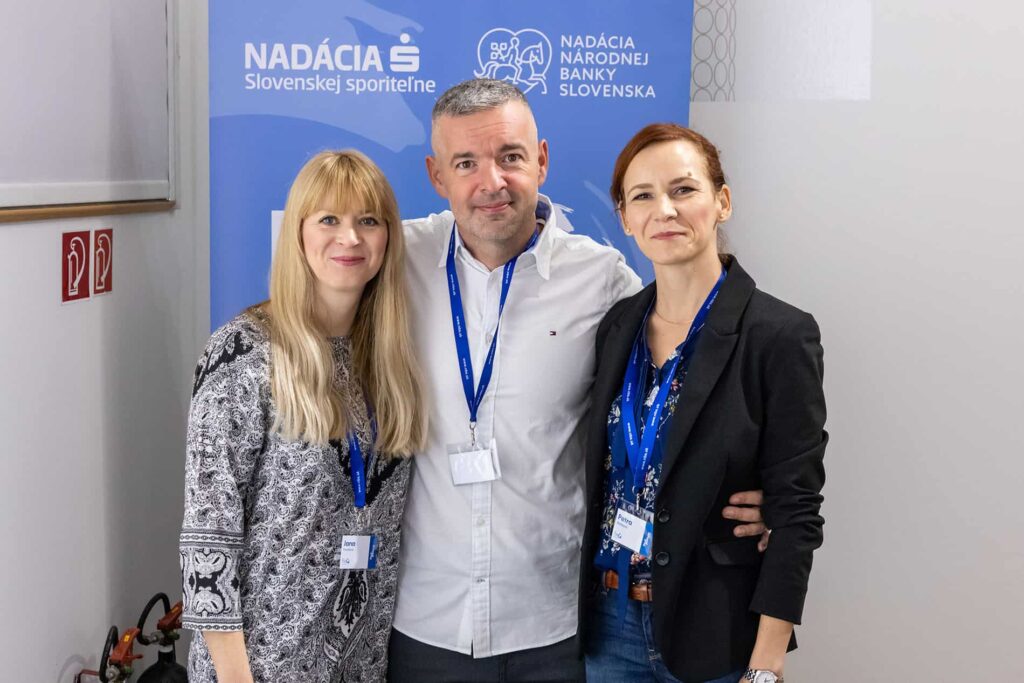 Traja dospelí s modrými šnúrkami stoja spolu a usmievajú sa do kamery. Za nimi je modrý transparent so slovenským textom a logami Nadácie Slovenskej sporiteľne a Nadácie Národnej banky Slovenska, oslavujúci Deň peňazí.